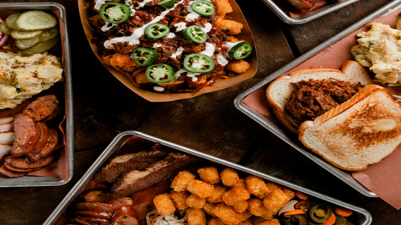 A tray of barbecue ribs with coleslaw, a dish of loaded tater tots with jalapeños and sour cream, and sides of pickles on a wooden table