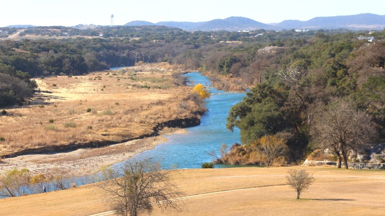 A river surrounding by green woodlands and brown fields near Pipe Creek, TX.