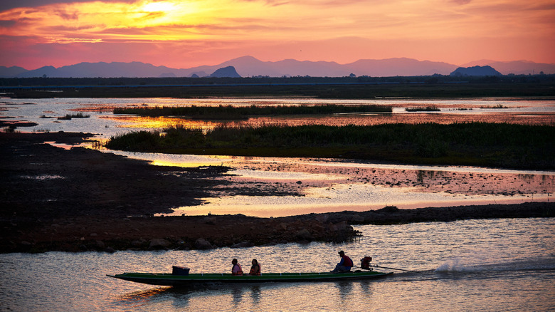 A long-tail boat with three passengers cuts across Songkhla Lake at sunset in Thailand.