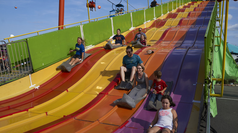 People of various ages slide down a rainbow-colored slide on rugs at The Great New York State Fair, Syracuse