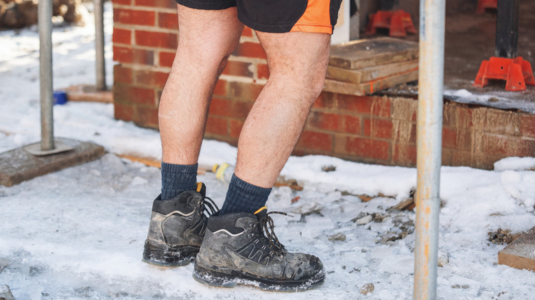 A person stands on a snowy site in shorts and work boots
