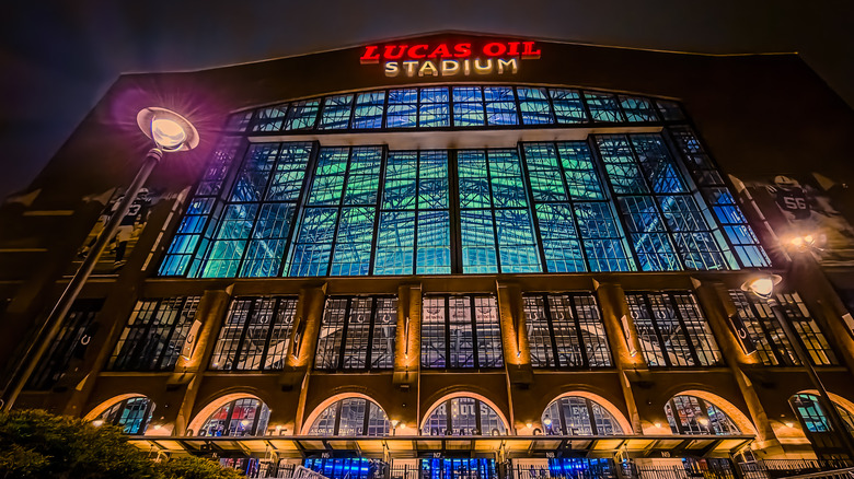 View of Lucas Oil Stadium entrance at night with retractable roof closed and blue and green lights lighting up the interior through glass windows