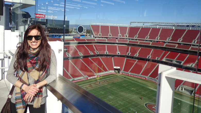 Brunette woman wearing sunglasses and a plaid blanket scarf on the rooftop deck of Levi's Stadium with the football field in the background on a sunny day