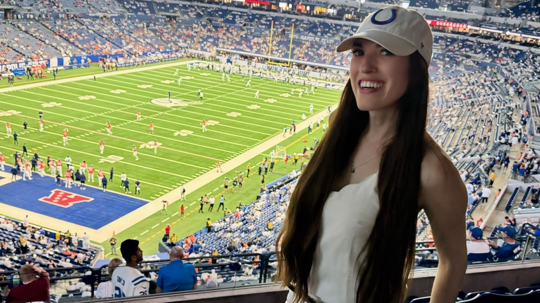 Woman with dark hair wearing a white Colts ball cap and a white outfit on Colts gameday with Lucas Oil Stadium football field in the background