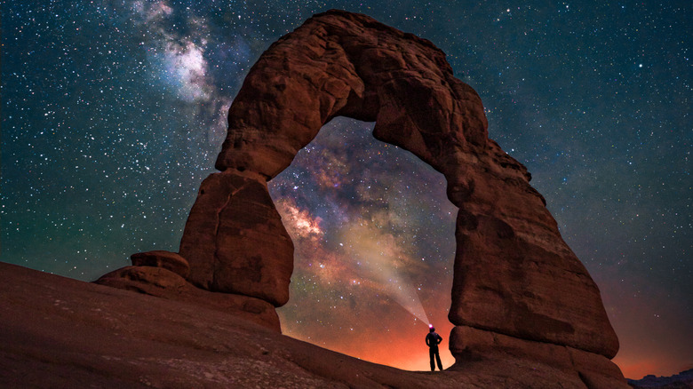 A photographer standing under the Delicate Arch with the Milky Way in the background.