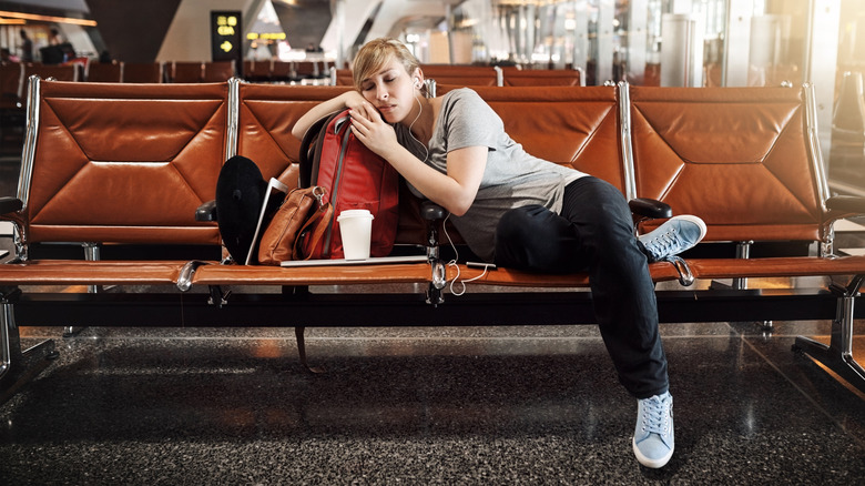 A passenger sitting in an airport terminal, leaning on their backpack with their eyes closed and earbuds in their ears