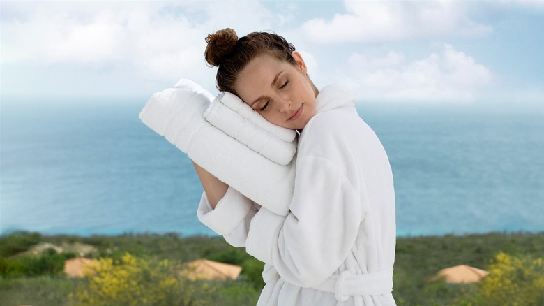 Woman in white robe hugging a set of soft white towels in front of ocean background