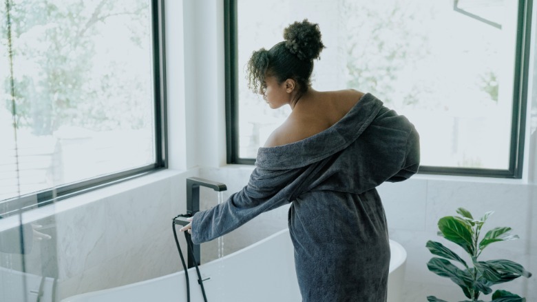 Woman in cozy gray robe turns bathtub water on
