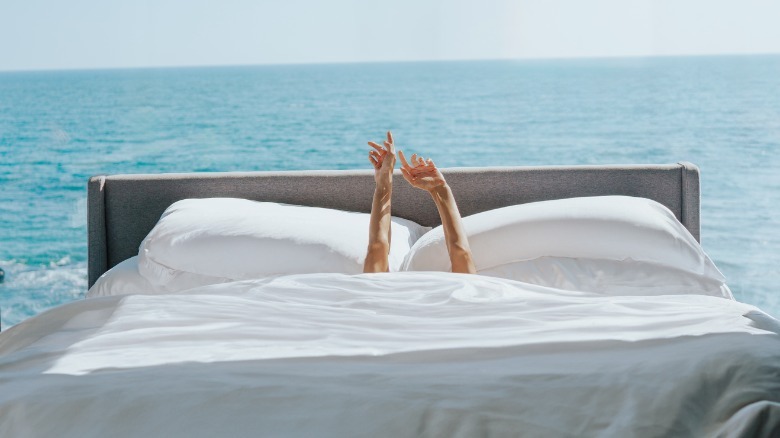 Hands reach out of bed surrounded by white bedding and an ocean backdrop