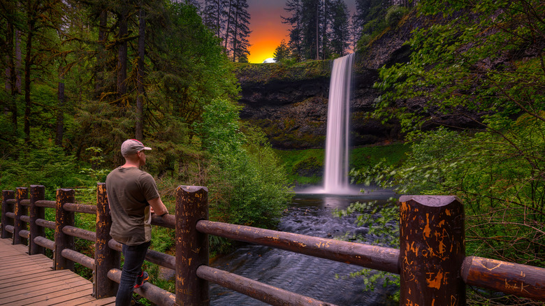 South Falls waterfall flowing over a rock cliff surrounded by trees at sunset, with a man on a wooden bridge in the foreground
