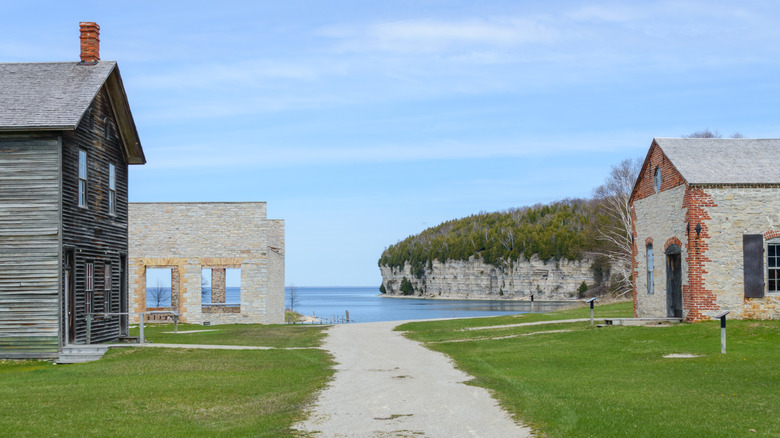 Fayette ghost town in Michigan, with abandoned buildings to either side of a path, with a lake in the background under a blue sky