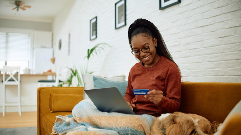 Woman smiling and shopping online while sitting on couch with dog