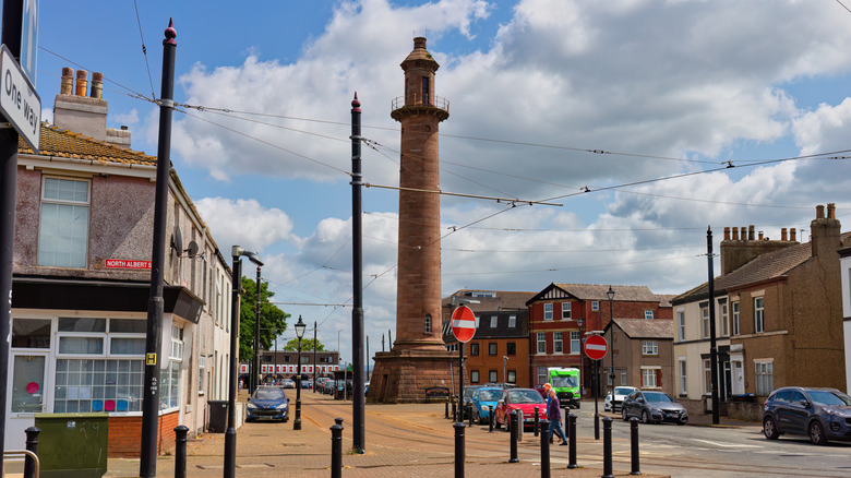 A tall, cylindrical monument stands in the center of a town square surrounded by buildings and parked cars under a partly cloudy sky in Fleetwood