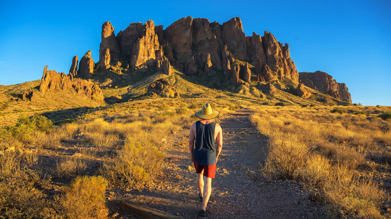 Hiker in a straw hat walking towards rock formations named Superstition Mountains in Lost Dutchman State Park, Arizona