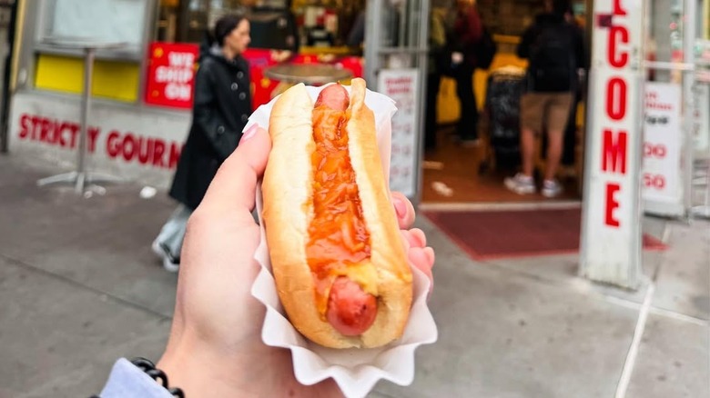 a hand holds up a hot dog in front of Gray's Papaya