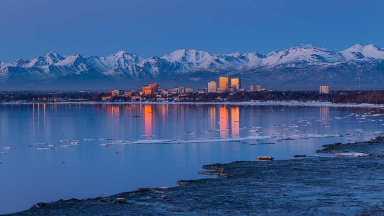 Lit-up buildings reflect in bay as snow-capped mountains rise in background of Anchorage, Alaska