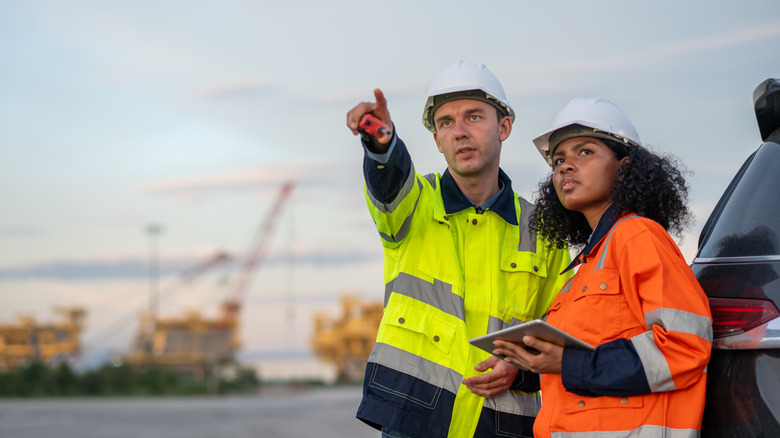 Man and woman in hard hats and hi-viz coats look on as cranes rise in the background