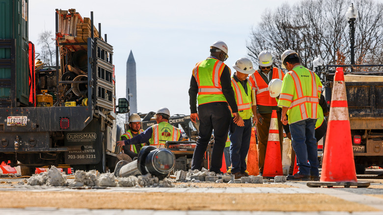 Workers stand around road construction on Washington, D.C., street as Washington Monument looms in background