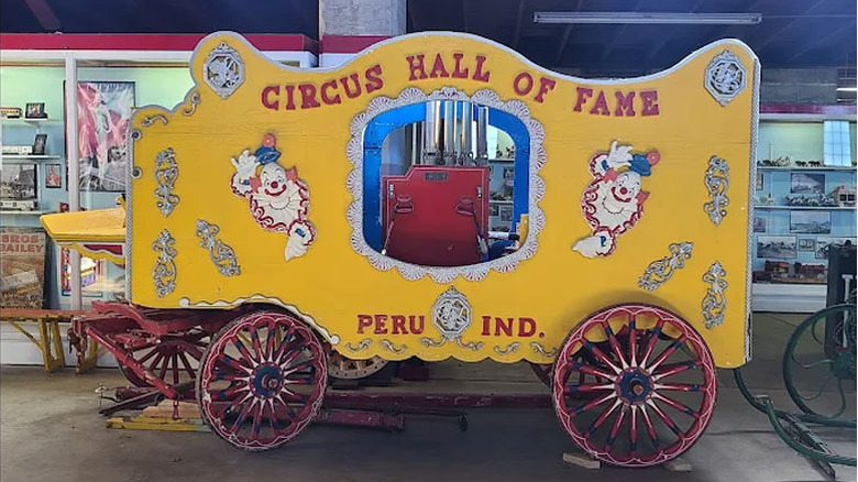 Yellow wagon with clown images and red wheels that reads Circus Hall of Fame Peru Ind., with display cases in the background