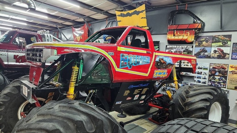 Red monster truck with yellow accents in front of a wall of monster truck photos and doors