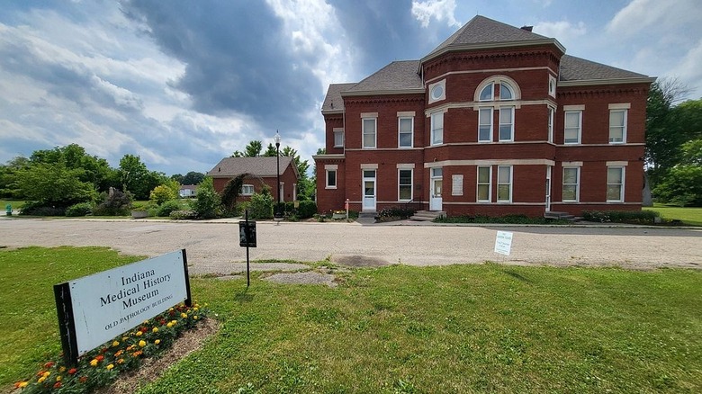 Red brick building with a sign by the parking lot reading Indiana Medical History Museum, surrounded by trees under a cloudy blue sky