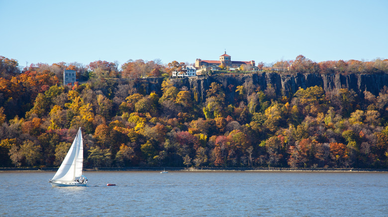 Sailboat on the water in front of a cliff covered in autumn foliage with homes built atop it, under a blue sky