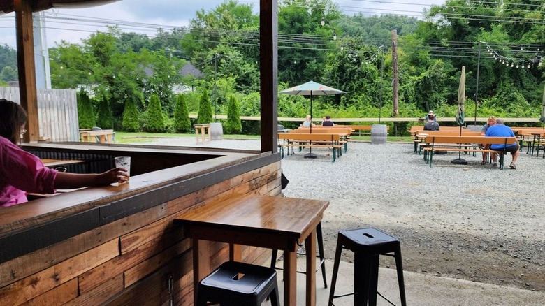 Wooden bar with a two-top table in the foreground, with wooden picnic tables in a gravel lot