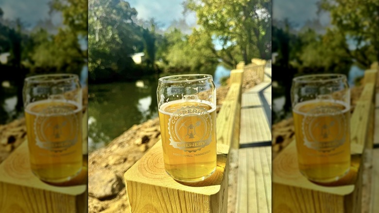 Yellow beer in Copperhill Brewery glass on a wooden railing, with a river and trees in the background, under a blue sky
