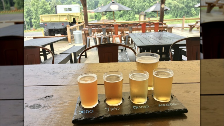 Flight of beer on a wooden table in front of chairs, picnic tables, black umbrellas, and a yellow truck, with green trees in the background