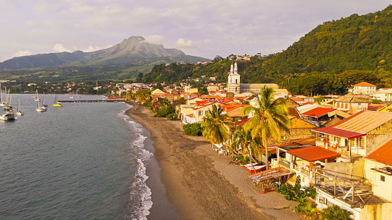 A charming coastal town in Dominica is featuring vibrant buildings along a sandy beach. Sailboats are anchored nearby on the calm sea, while lush green mountains are rising in the background under a partly cloudy sky