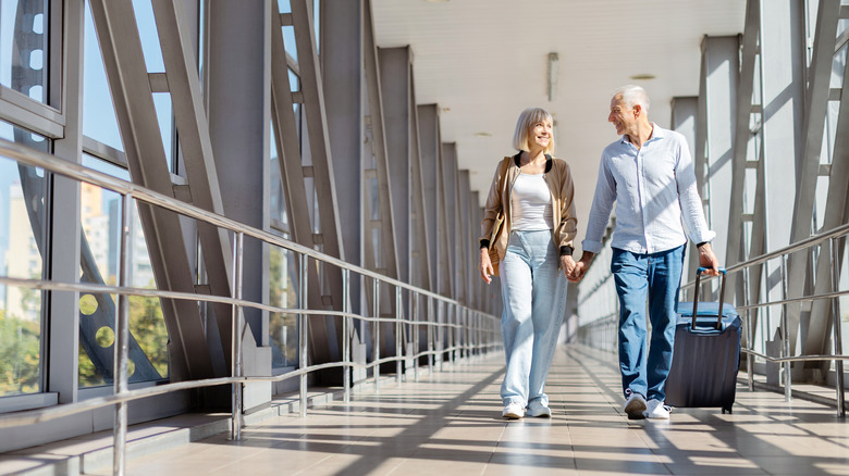 Happy senior couple walking at airport corridor with luggage, holding hands