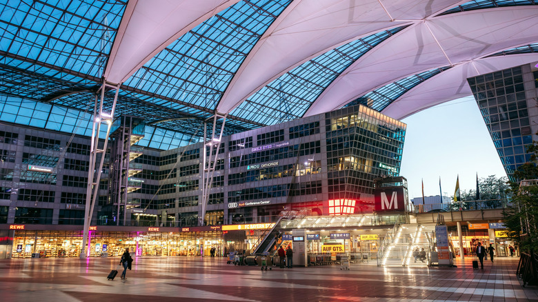 Interior plaza of Munich Airport with futuristic glass roof, illuminated staircases, shops and passengers walking in the evening