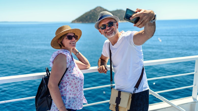 Middle-aged couple taking a selfie leaning on a cruise ship rail with blue water and an island in the background