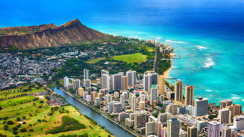 Aerial view of Honolulu facing the deep blue ocean and framing its skyscrapers, green spaces, canal, and Diamond Head crater in the distance.