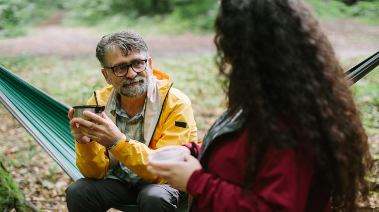 A man sitting in a hammock while camping, drinking tea with a woman.