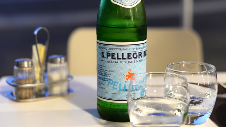 A bottle of San Pellegrino stands opened before two glasses of water on a white linen table with salt and pepper
