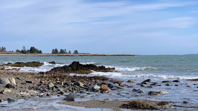 A pale blue ocean crashes up against rugged brown and gray rocks with a big blue sky full of cirrostratus clouds at The Anchorage Provincial Park in New Brunswick, Canada