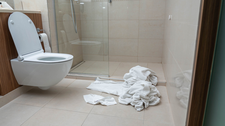 Bunched-up towels lie on the floor of a tiled bathroom in front of an open toilet