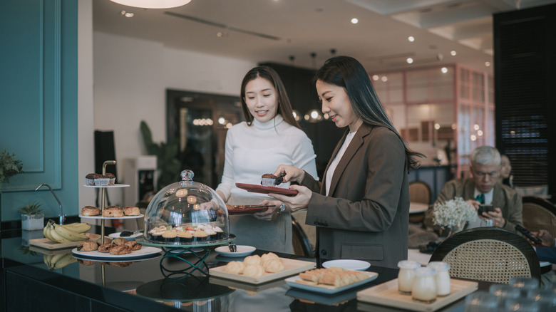 Two well-dressed young women serving themselves pastries at an airport lounge buffet table