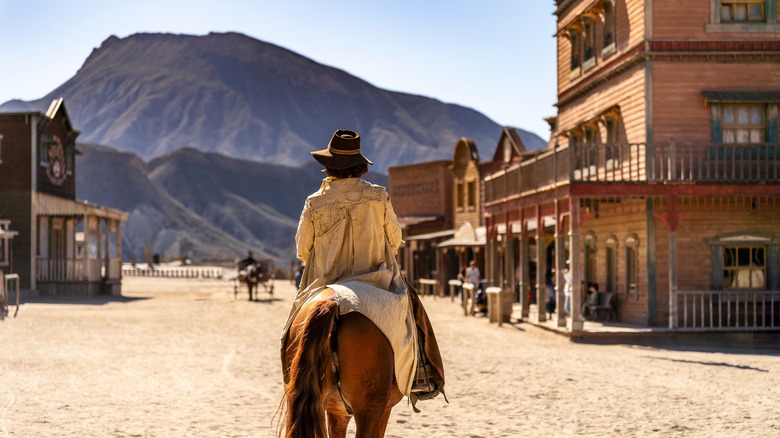 Rider with coat and hat atop a chestnut horse flanked by wooden western-style buildings and a mountain