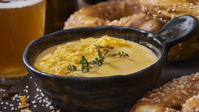 Individual crock of beer cheese sauce with an herb sprig on top and soft pretzels and beer in the background