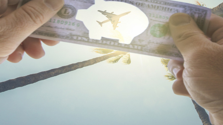 A dollar bill with a piggy bank-shaped cutout held up to the sky as a plane flies overhead.