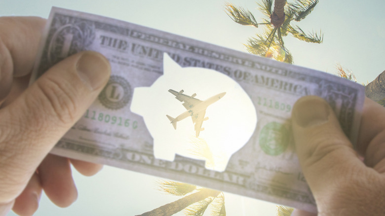A dollar bill with a piggy bank-shaped cutout held up to the sky as a plane flies overhead.