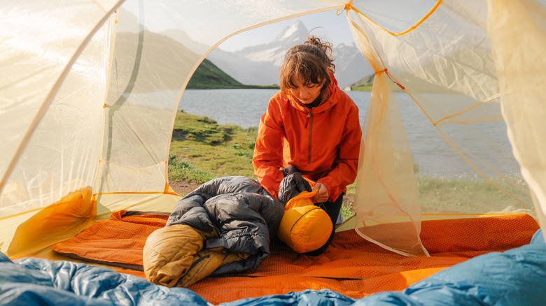 woman stuffing a sleeping bag into a bag in a tent beside a mountain lake