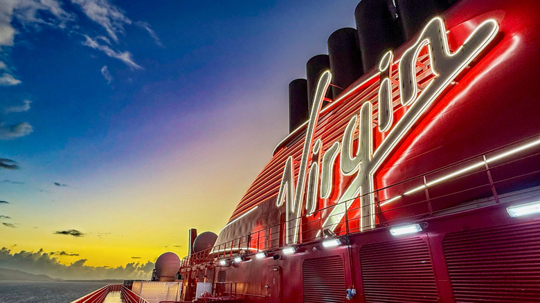 Lighted Virgin logo on the side of a red Virgin Voyages ship at sunrise
