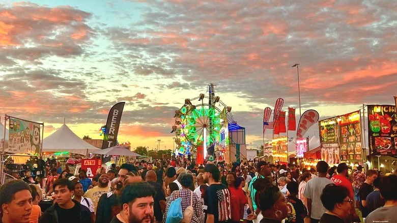People milling around vendors and park rides under an orange and red sunset at the Great American Foodie Festival
