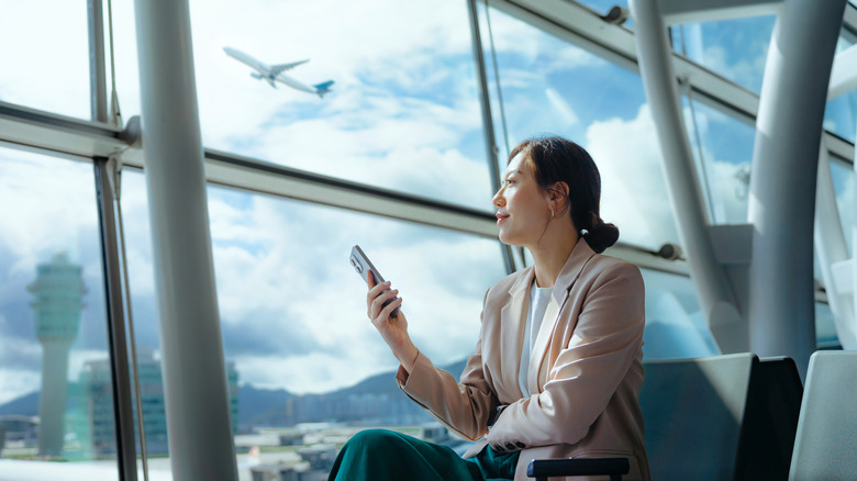 Woman staring out the window at an airport terminal with her phone in her hand and an airplane in the background