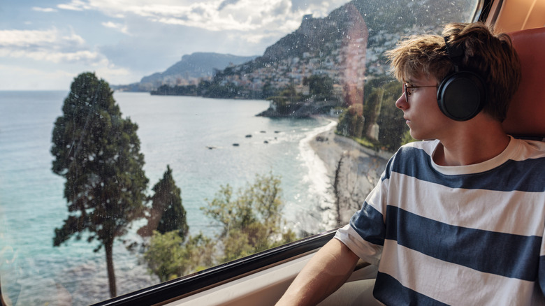 Teenager with earphones looking out a train window at the water.