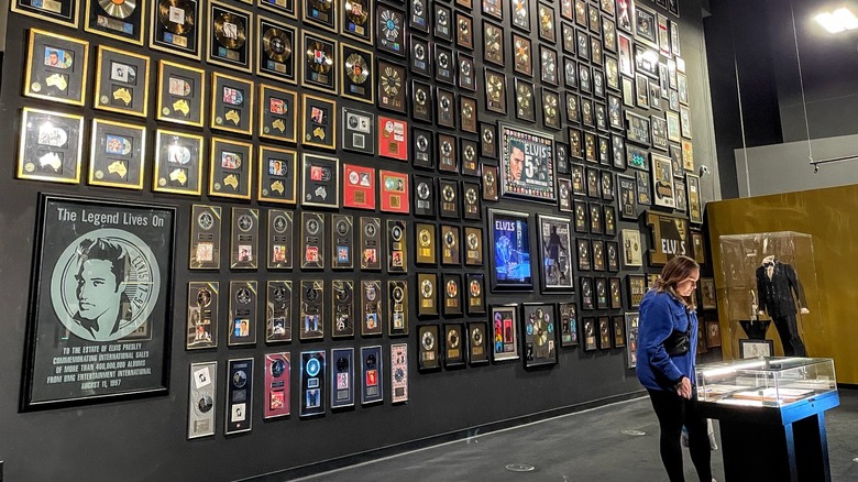 Elvis Presley museum exhibit with tall wall displaying gold records, woman looking at artifacts