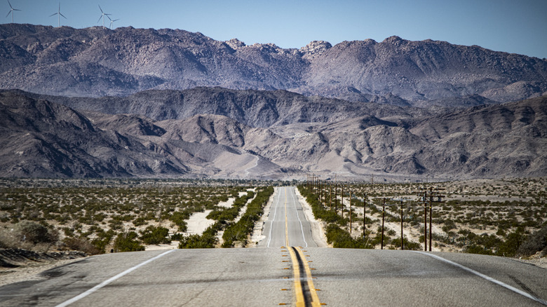 A long straight highway cuts through the middle of a desert with rocky mountains in the backdrop